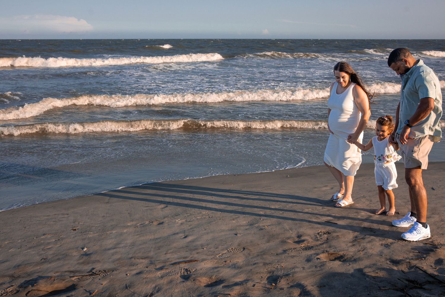 Family of three walking on a sandy beach near ocean waves at sunset, parents holding child's hands.