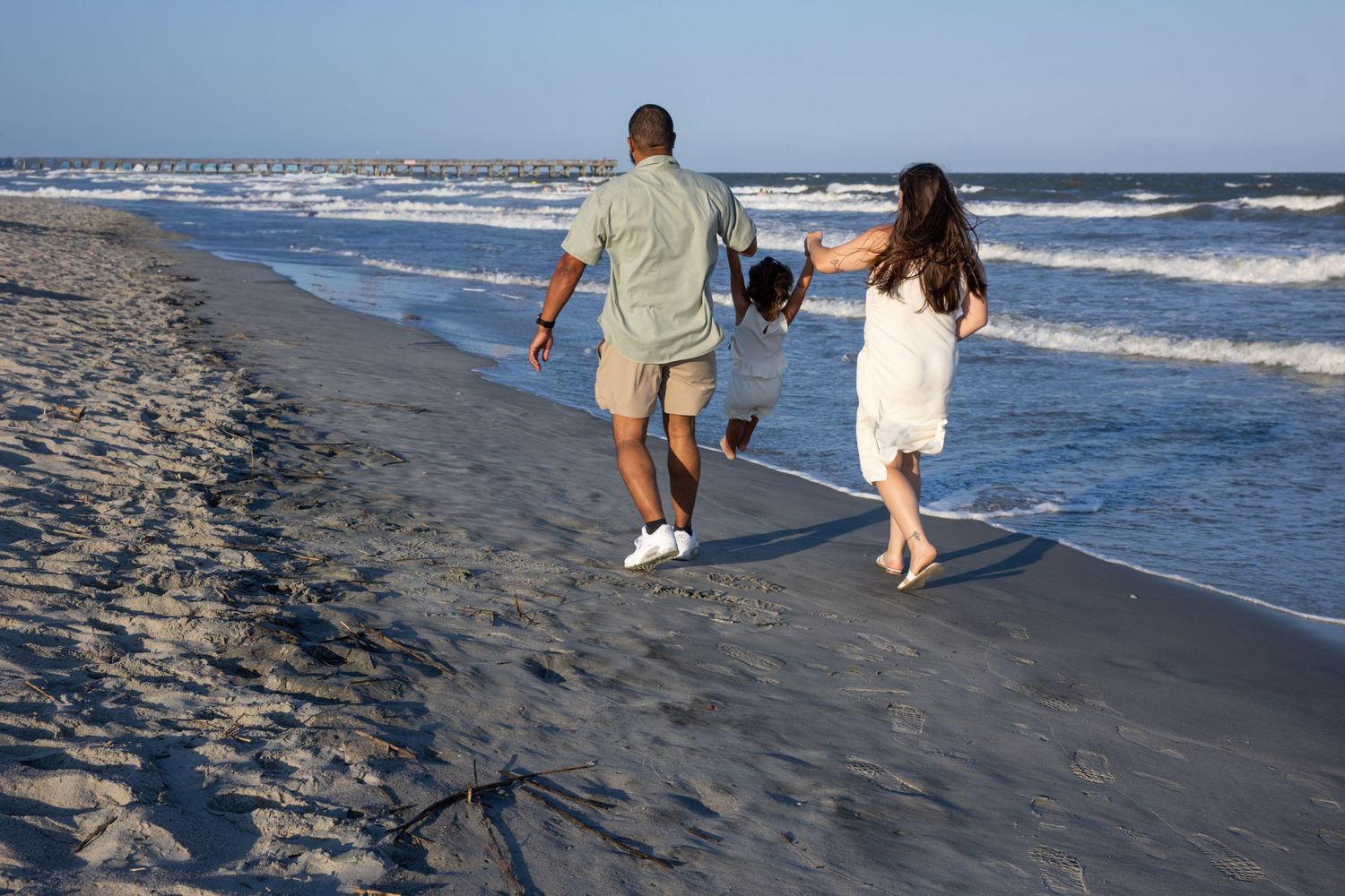 Family of three walking along a sandy beach shoreline, parents swinging child between them on a sunny day.