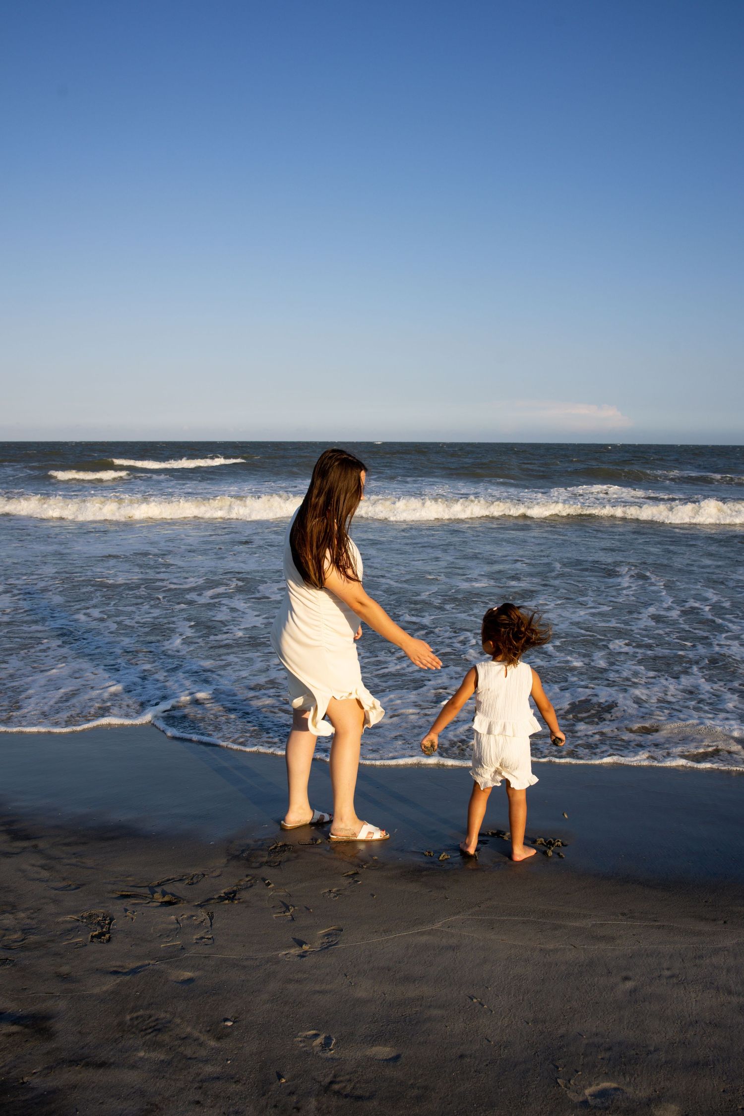Two girls in white dresses play at ocean's edge on a sandy beach with gentle waves under a clear blue sky.