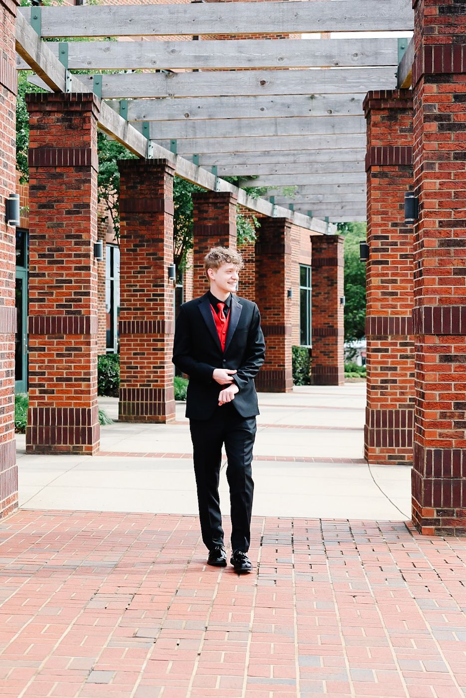 Young man in black suit with red tie walking through a brick pergola pathway.