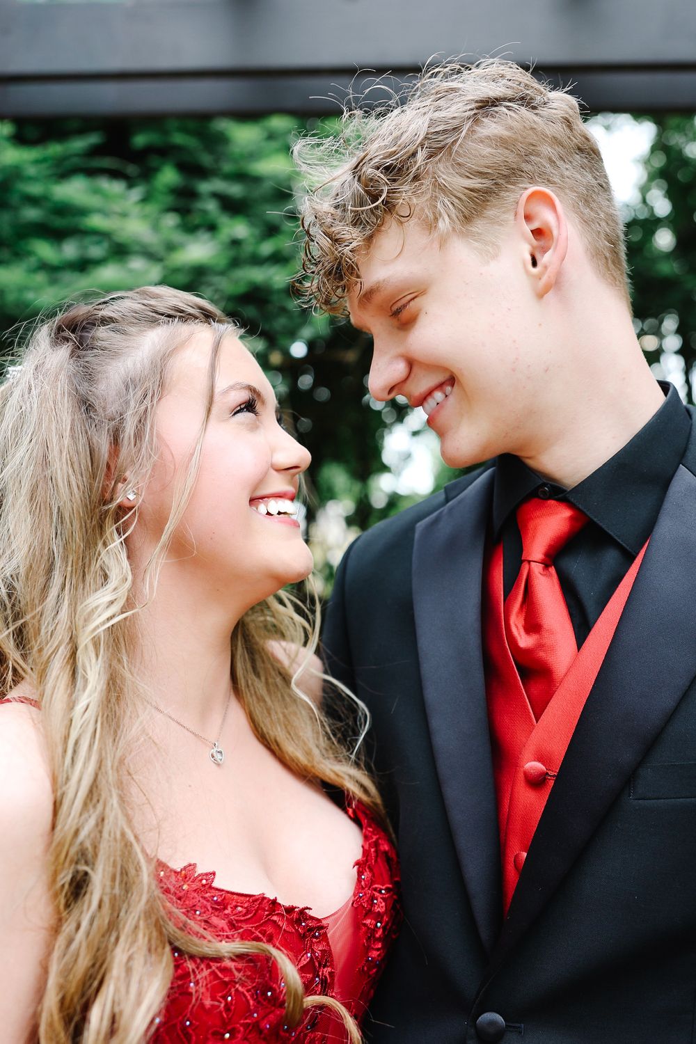 A happy teenage couple in coordinating red and black prom outfits smile at each other outdoors.