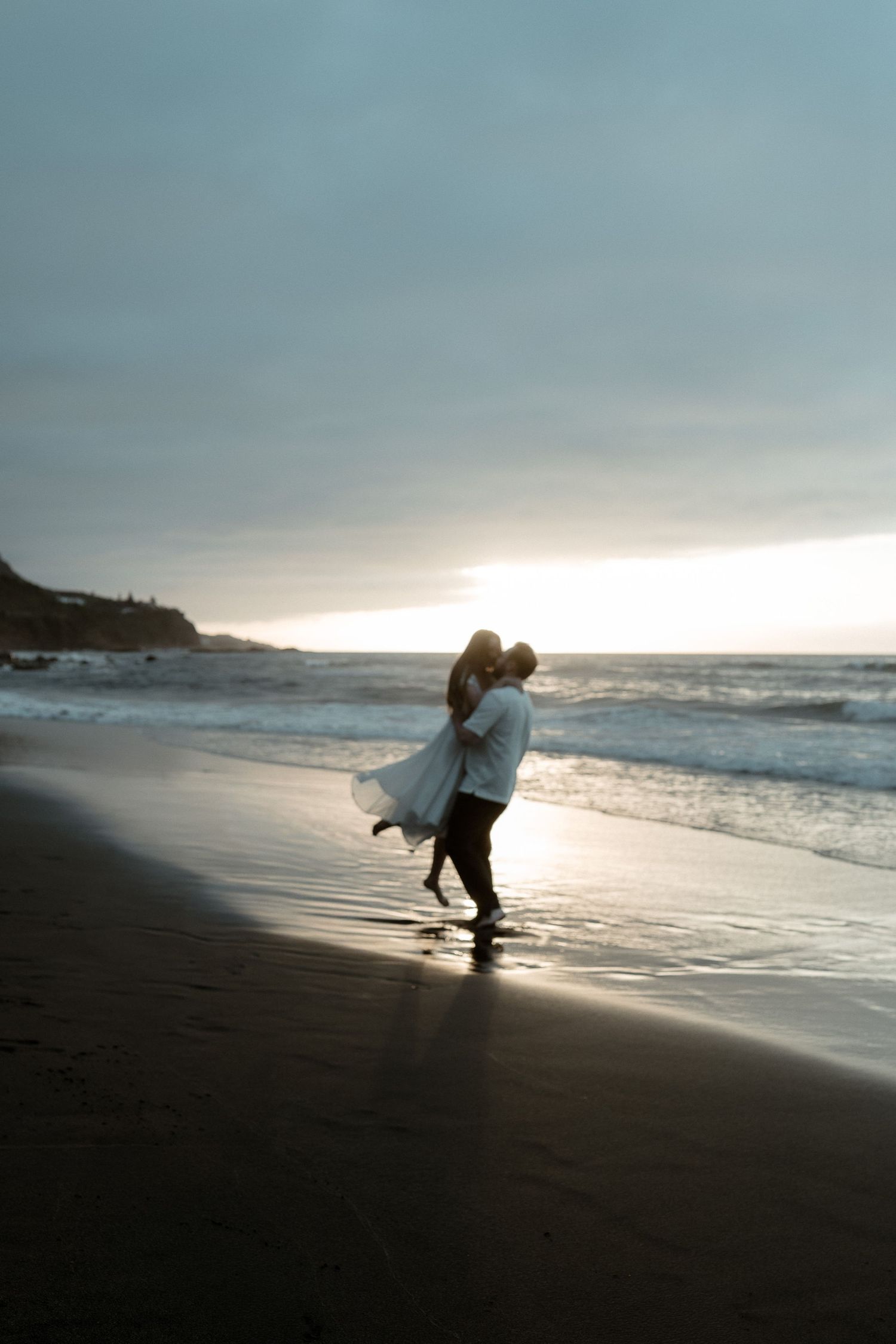 A couple shares a romantic embrace on a sandy beach at sunset, with ocean waves and dramatic cloudy sky behind them.