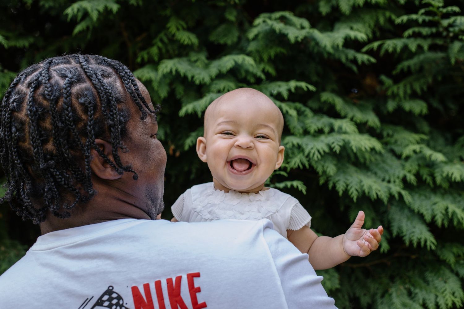 A joyful baby laughing while being held up by a person wearing a Nike shirt outdoors near green trees.