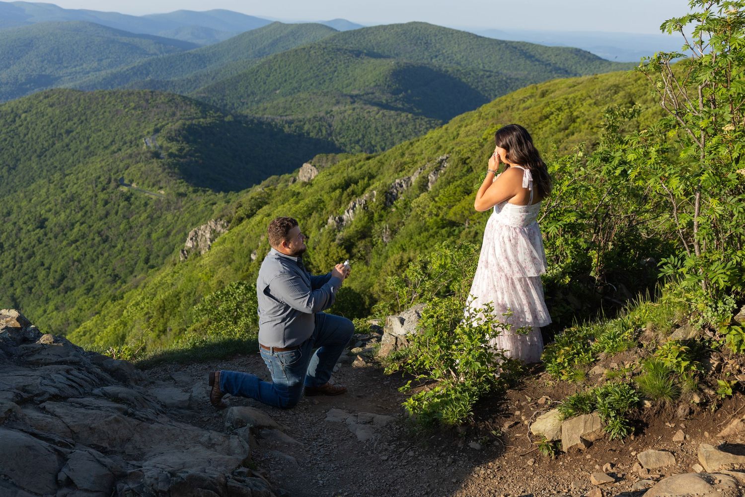 Man proposes on one knee to woman on scenic mountain overlook at Stony Man Mountain Summit surrounded by lush green hills before sunset.