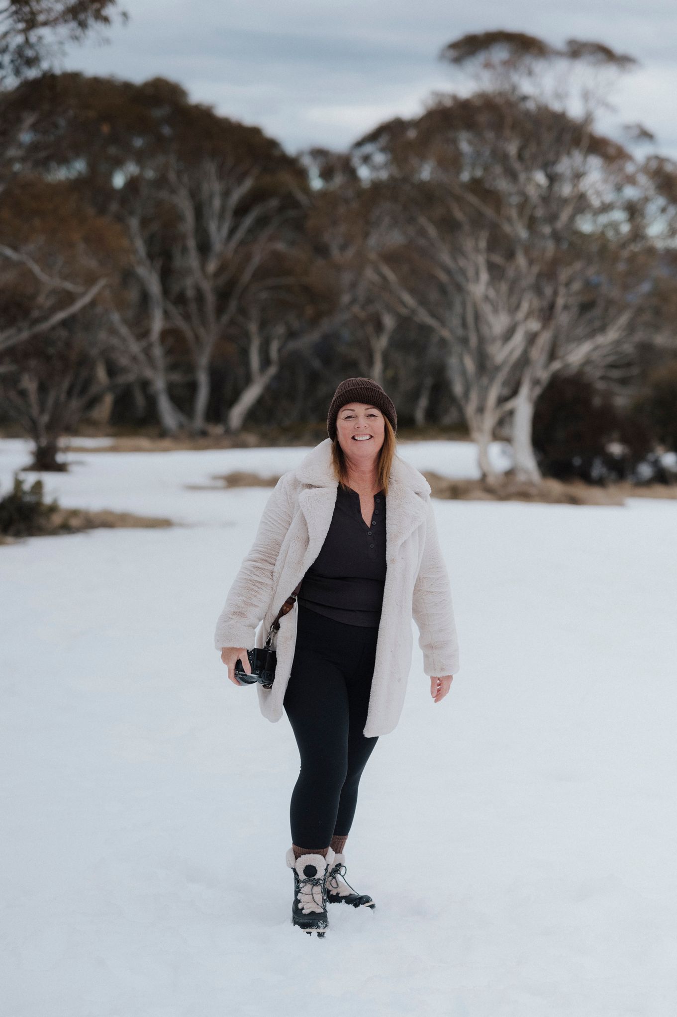 Woman in white fluffy coat and black leggings smiling while walking in snowy winter landscape with bare trees.