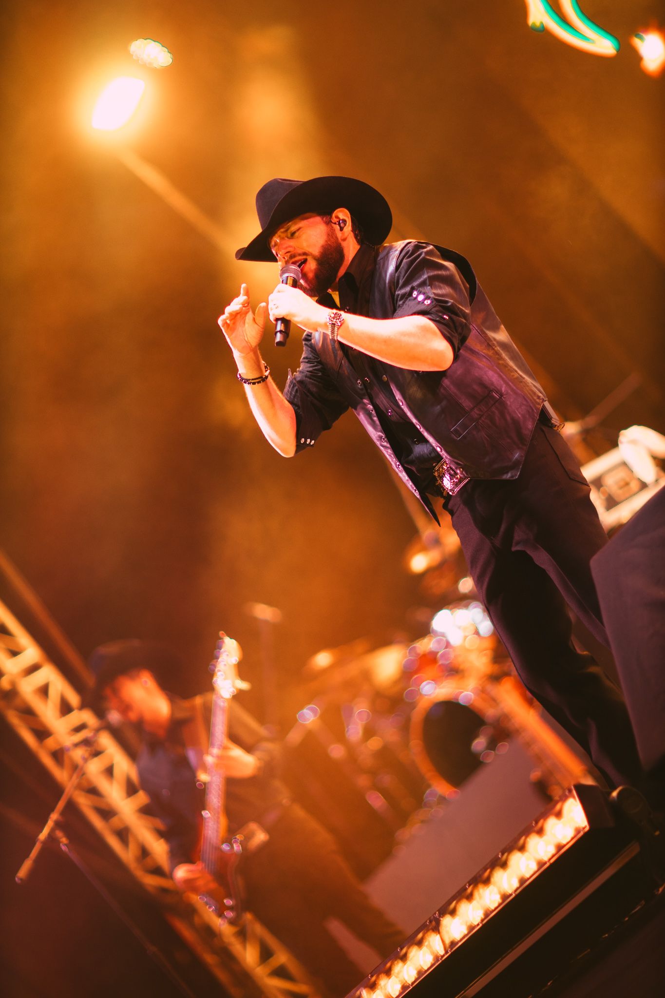 Male singer in black hat performing on stage under warm orange lights with band members visible in background.