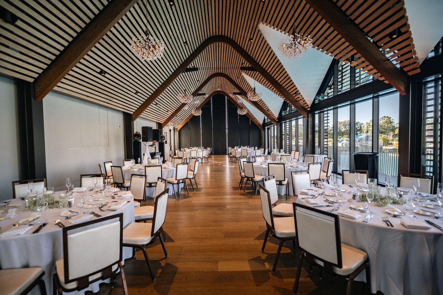 Elegant wedding reception hall with vaulted wooden ceiling, round tables set with white linens, and floor-to-ceiling windows.