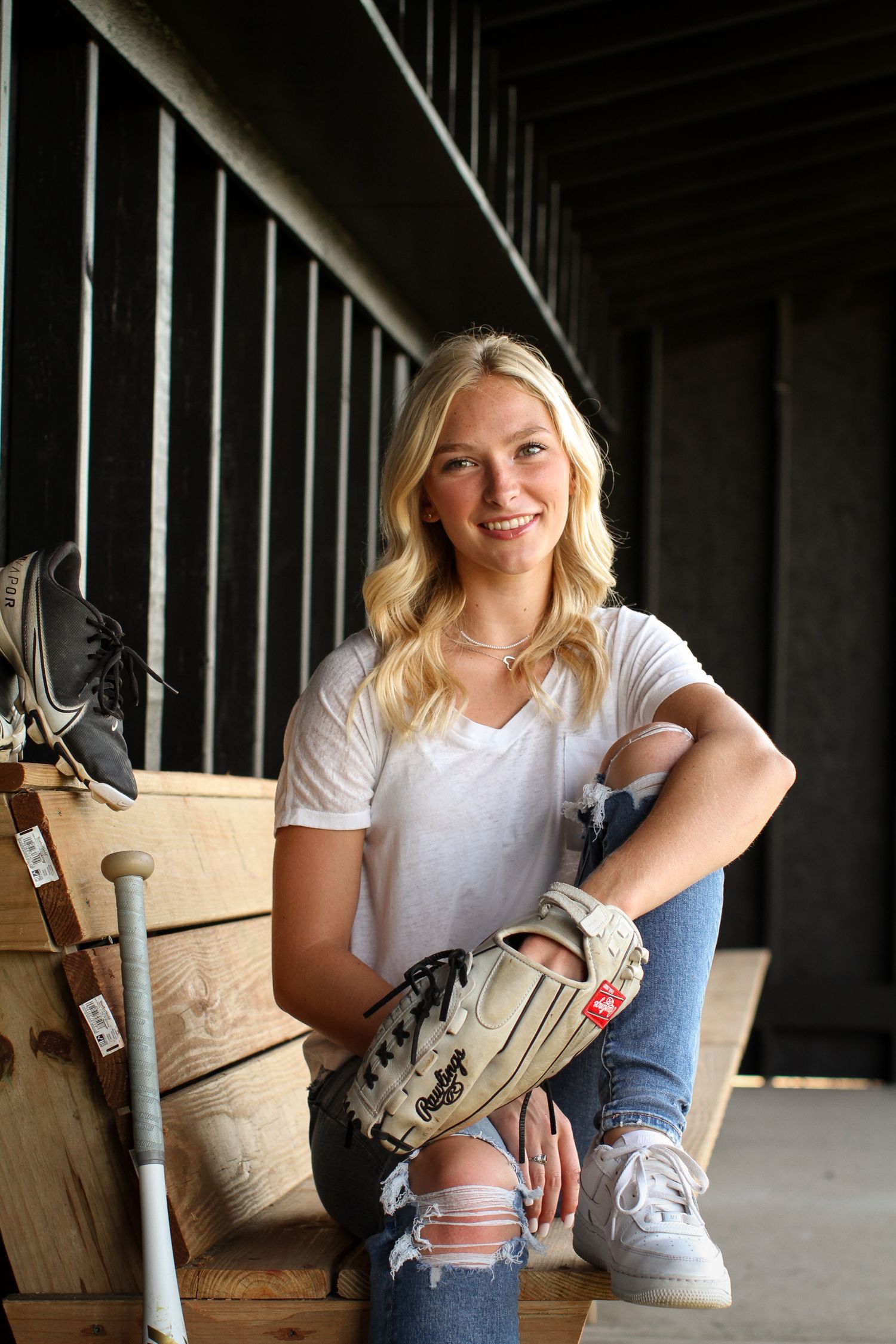 A young woman sits on the bench in a dugout and smiles at the camera while wearing a softball glove, her cleats and bat resting beside her.