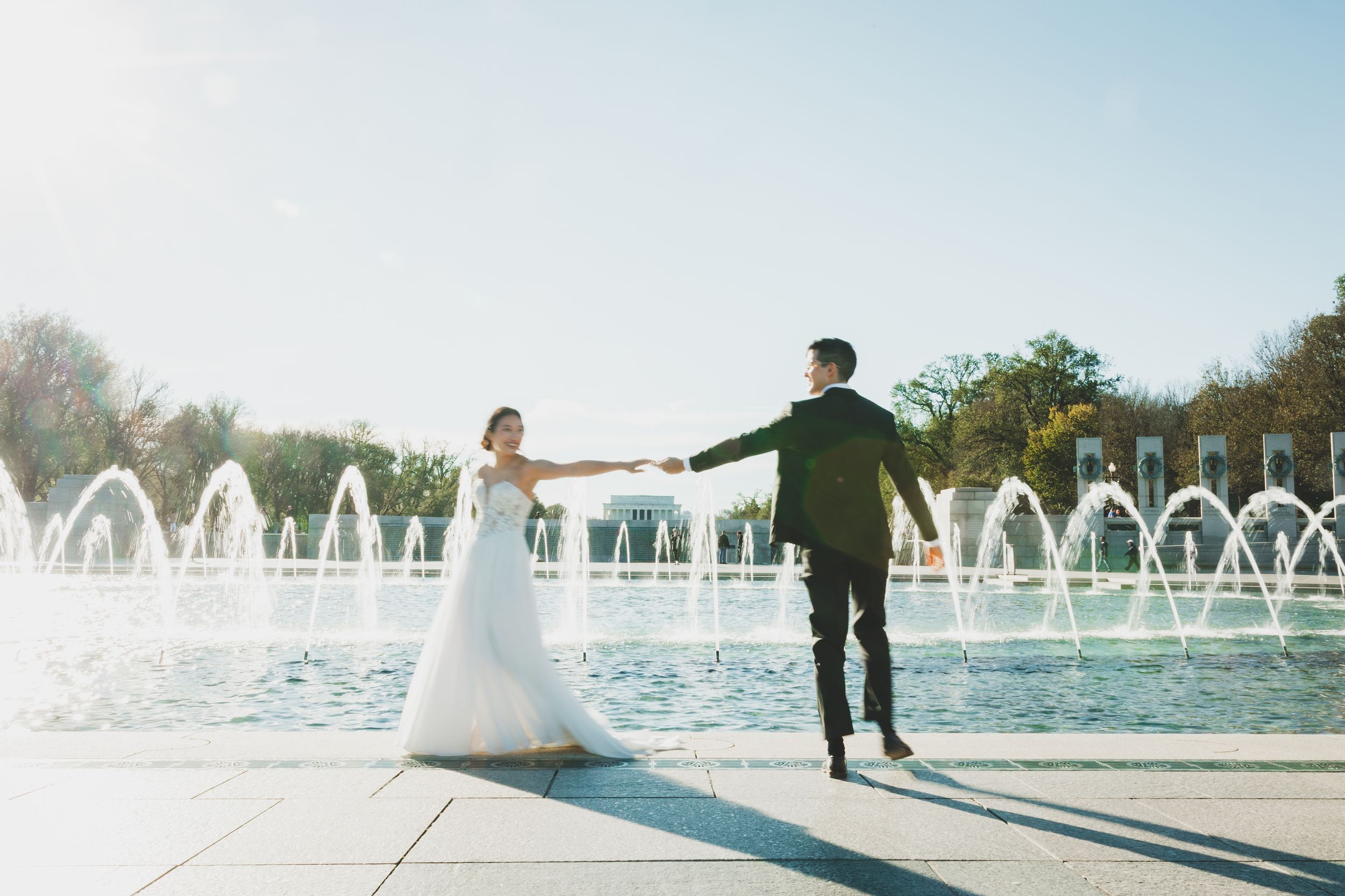 Bride and groom hold hands by fountain at WWII Memorial on sunny wedding day.