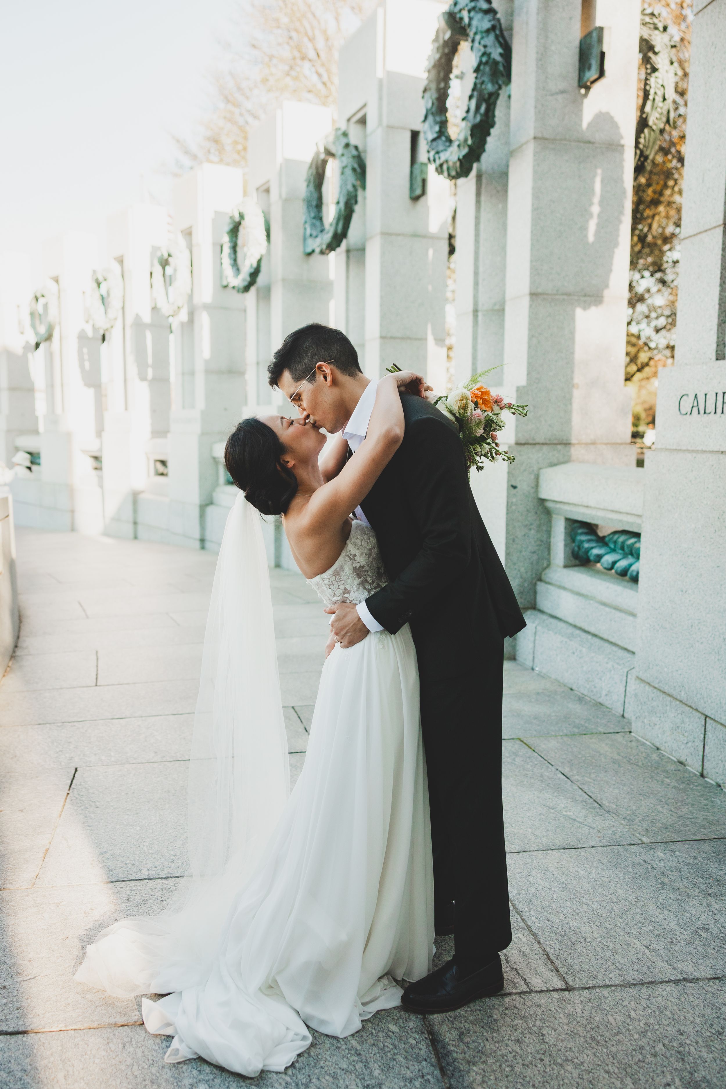 Bride and groom share a romantic kiss at the WWII Memorial in Washington DC, adorned with wreaths.