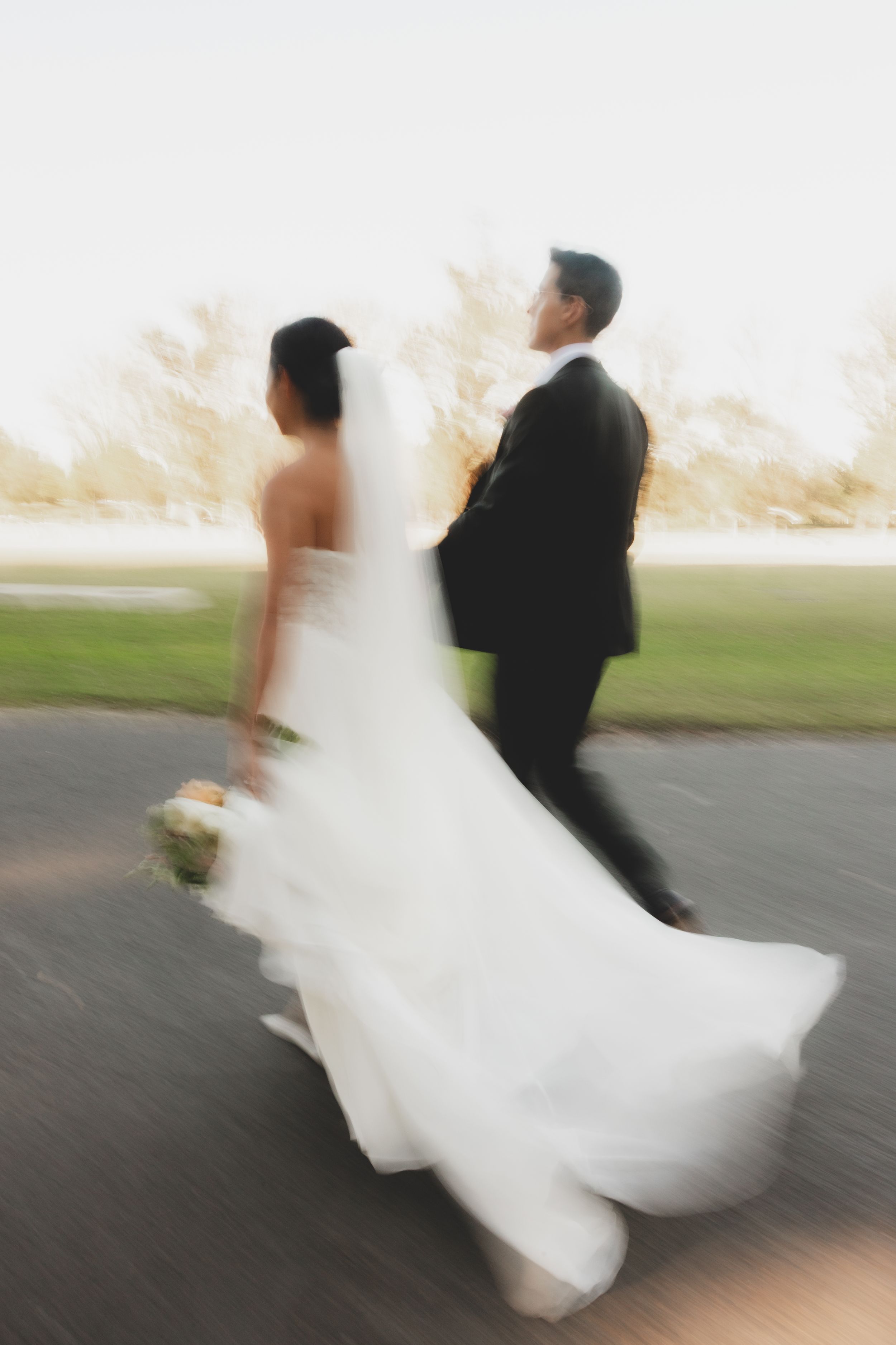 Blurred motion shot of bride in white gown and groom in black tuxedo walking together on their wedding day.