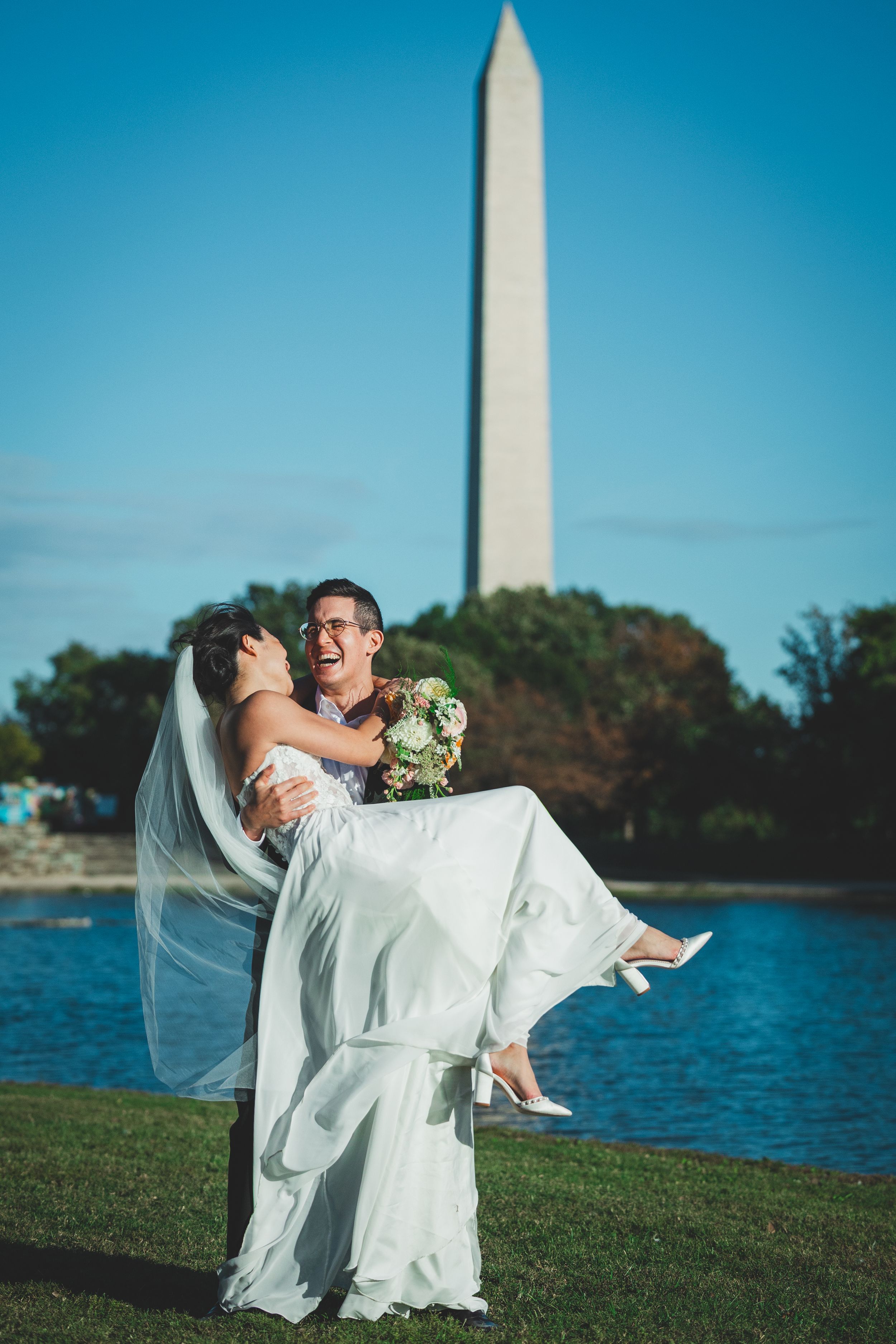Groom carries bride in white dress near Washington Monument reflecting pool on sunny wedding day.