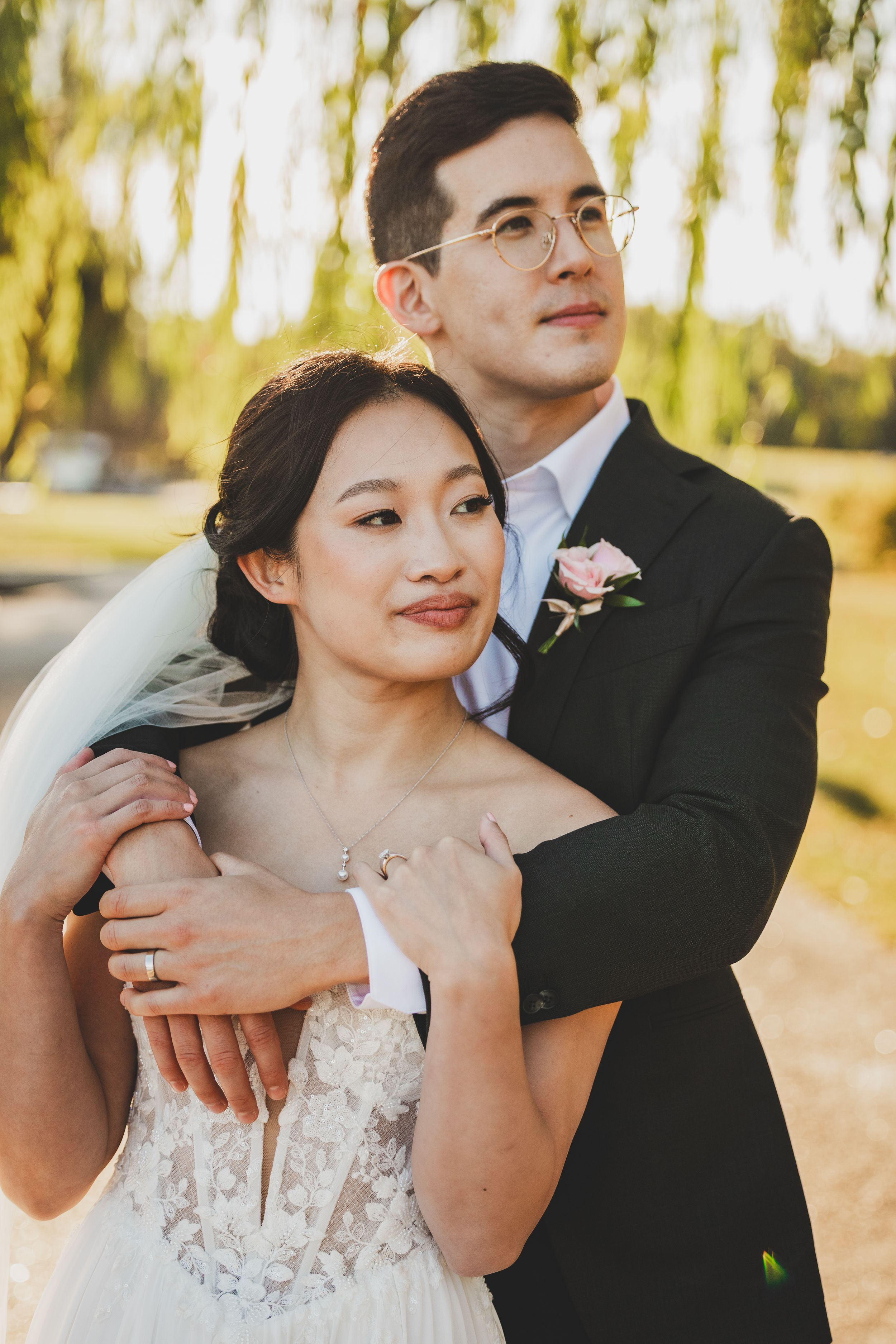 A bride and groom share a tender embrace outdoors, with the groom in a dark suit and bride in a lace wedding gown.