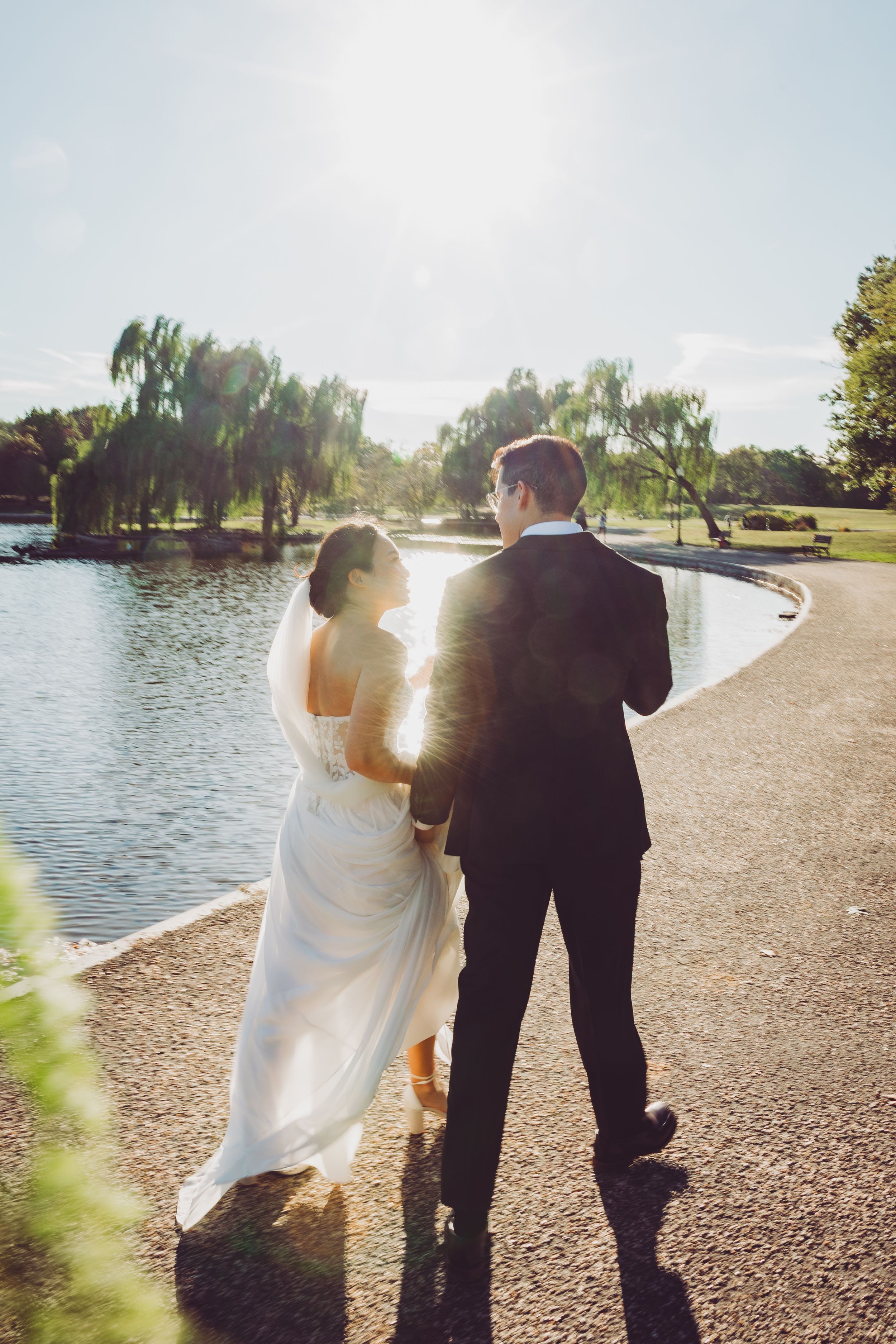 Bride and groom walking hand in hand along a scenic lakeside path surrounded by willow trees on their wedding day.