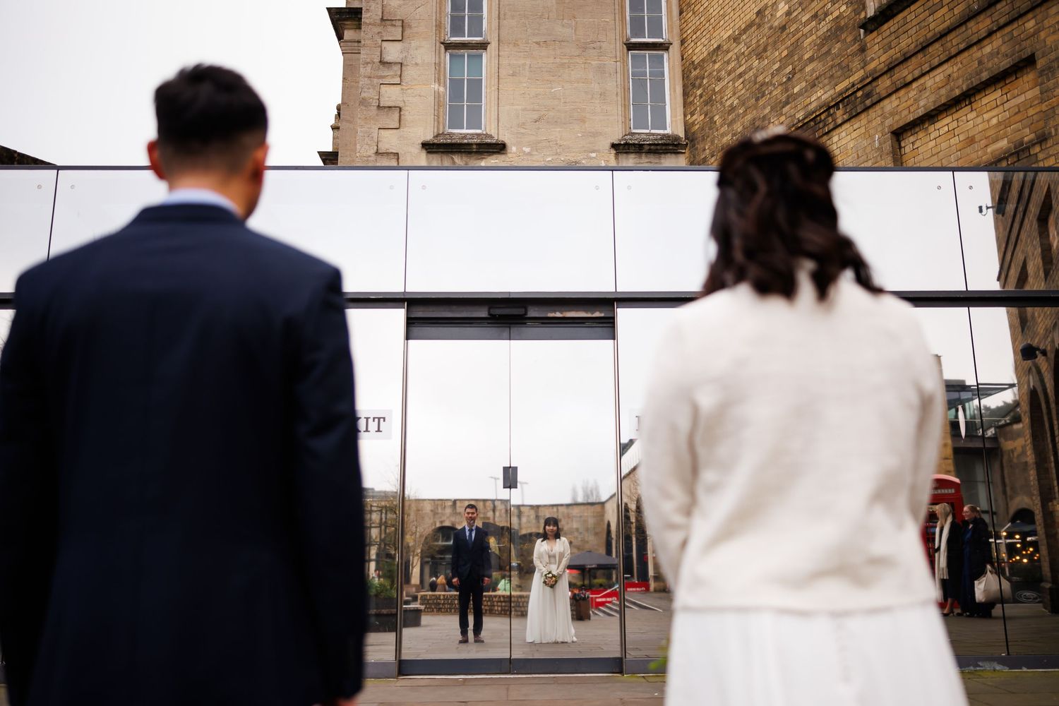 Couple stands before mirrored wall reflecting their wedding attire at an outdoor urban venue.