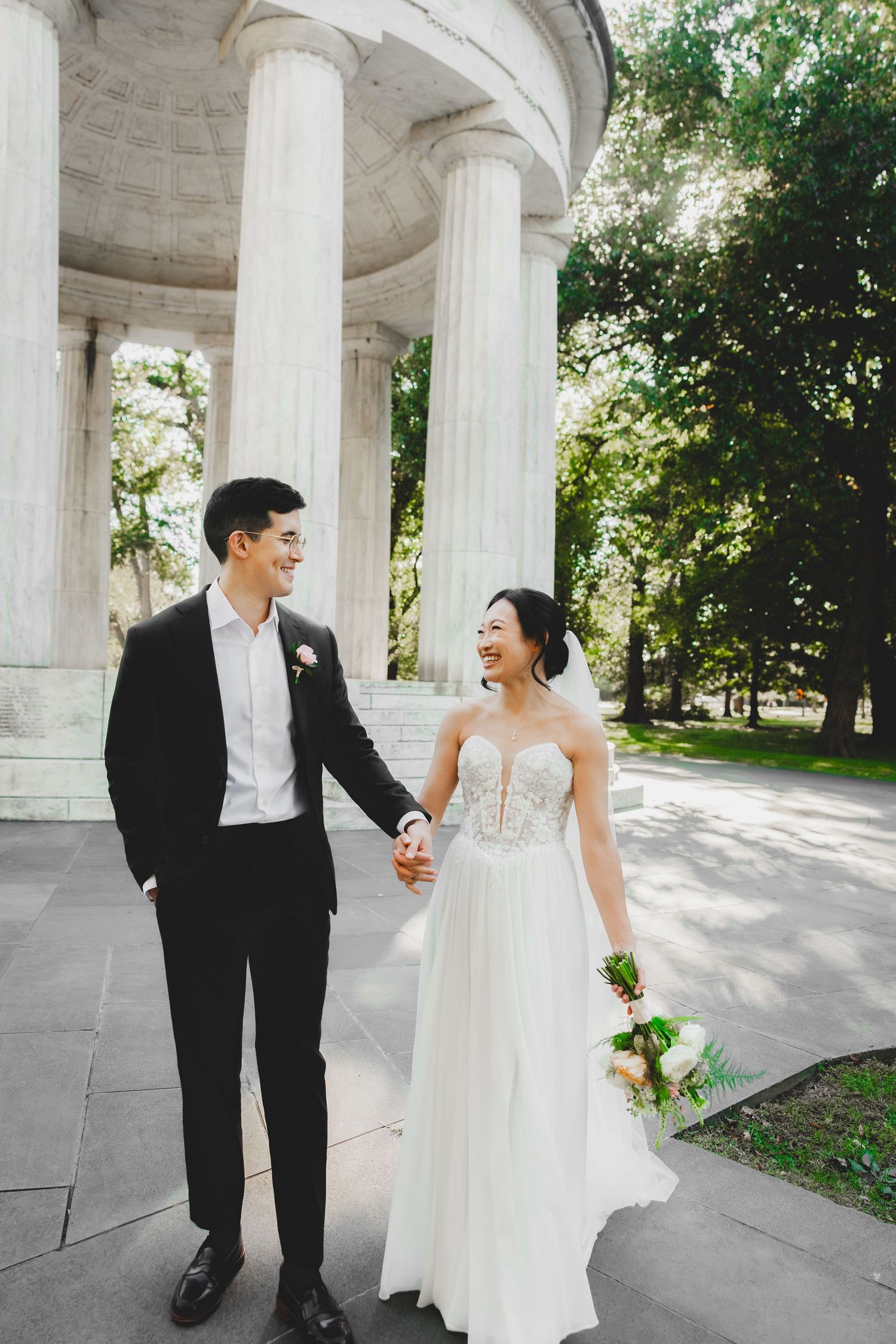 A smiling  bride and groom hold hands walking in front of the dc war memorial washington dc
