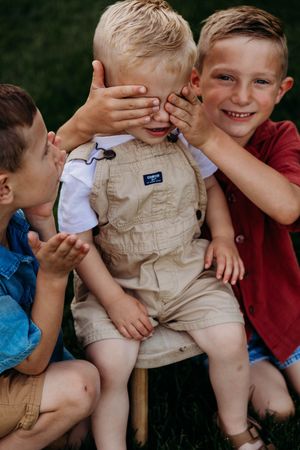 Three young boys playing together outdoors, with older siblings covering toddler's eyes in OshKosh overalls.