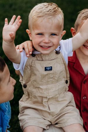 Smiling toddler boy in tan OshKosh overalls waving outdoors, surrounded by other children in a candid family photo.