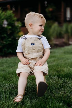 Toddler boy in tan OshKosh overalls and white shirt sitting on green grass outdoors, smiling softly.