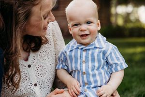 A smiling baby boy in a blue striped shirt being held by his mother outdoors in a green garden setting.