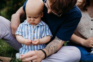 Adult with tattoo sitting on grass holding a baby dressed in blue striped outfit outdoors.