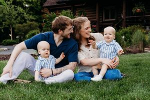 Happy family sitting on green grass with two baby twins in matching striped outfits outside a log cabin.