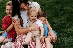 A mother hugs her three young children while sitting on a picnic blanket in a lush green grass yard.