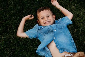A happy young boy flexes his muscles while lying on green grass, wearing a blue denim shirt.
