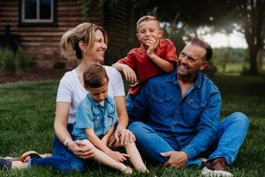 Happy family of four sitting on green grass laughing together outside a rustic wooden home.