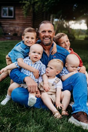 Smiling man sitting on grass with five young boys climbing on him near a wooden building.