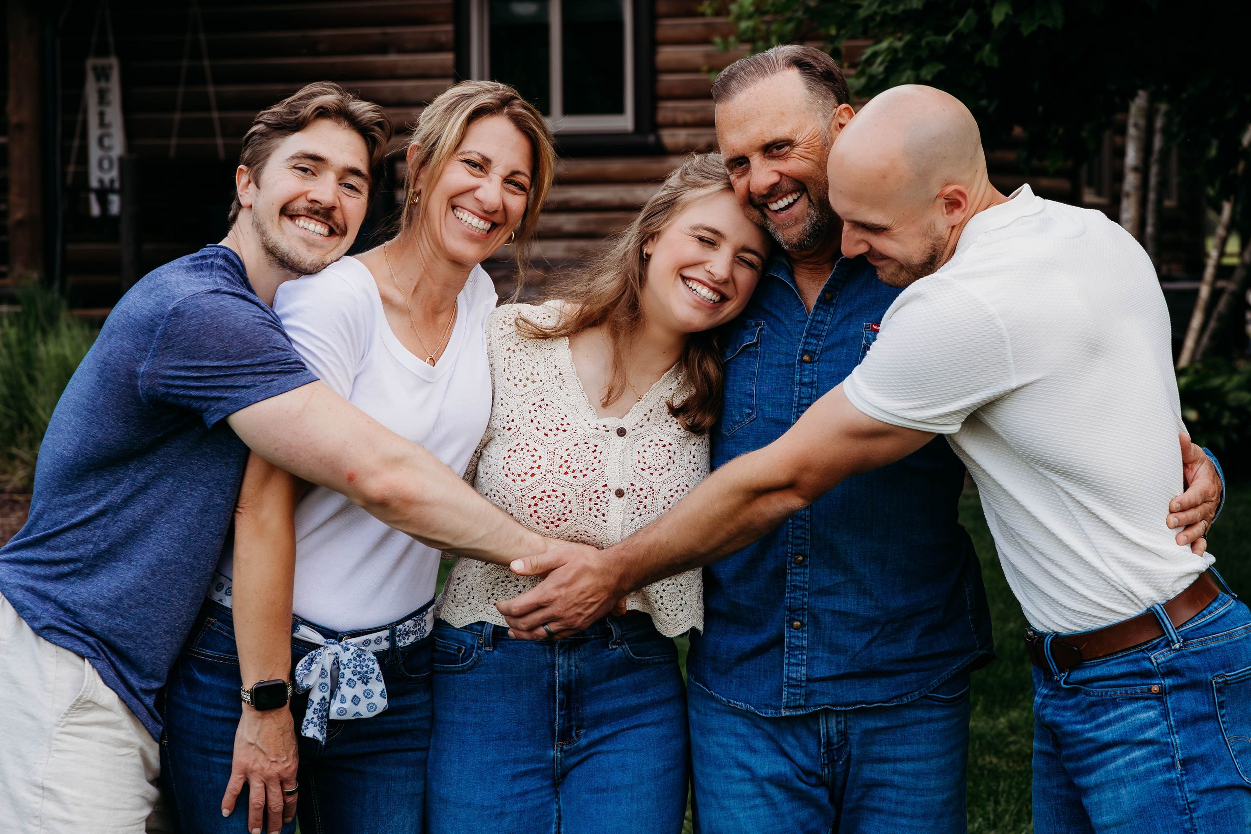 Happy family of five hugging and laughing together outdoors near a rustic wooden cabin.