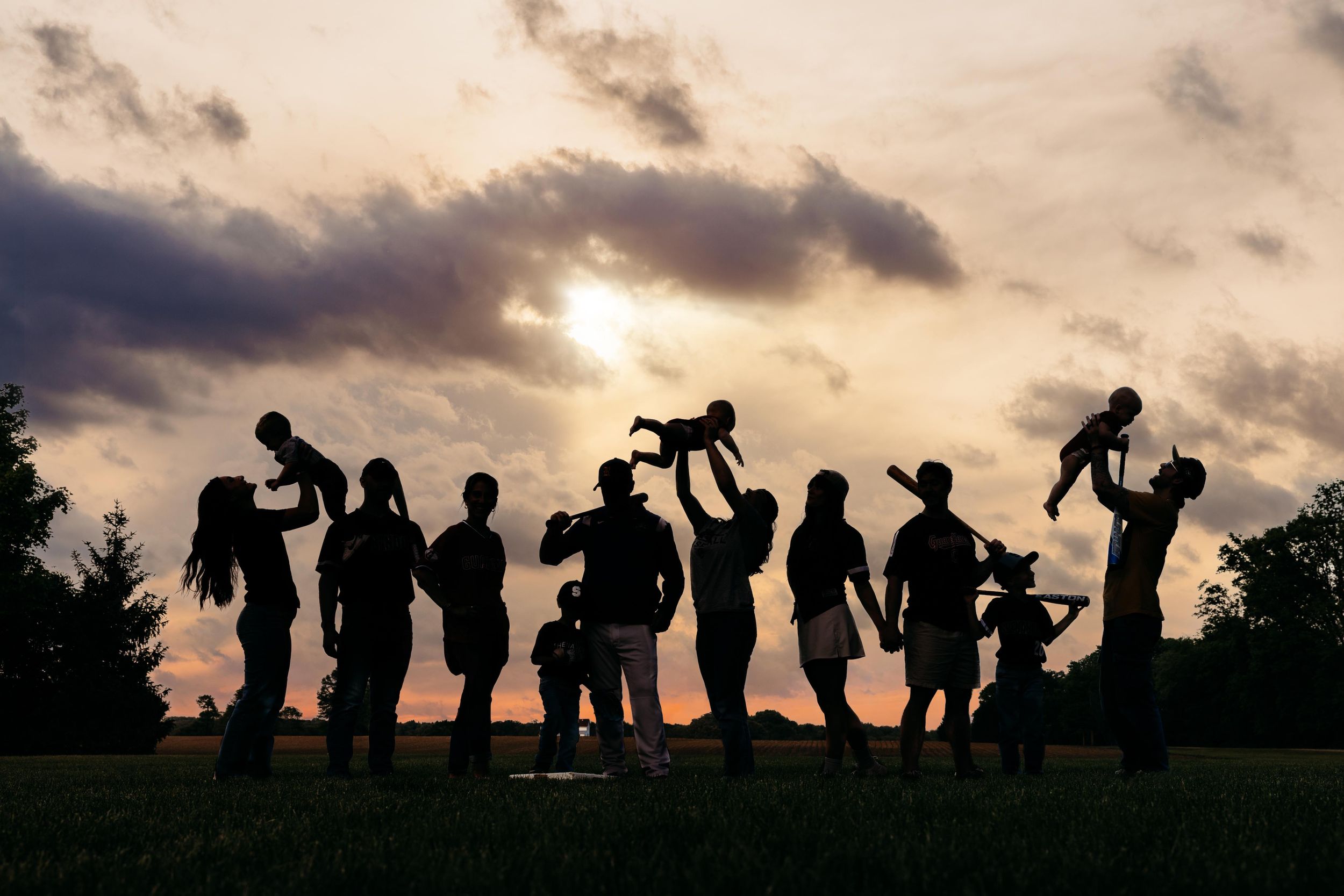 Silhouettes of a large family group lifting children at sunset against a dramatic cloudy sky in an open field.