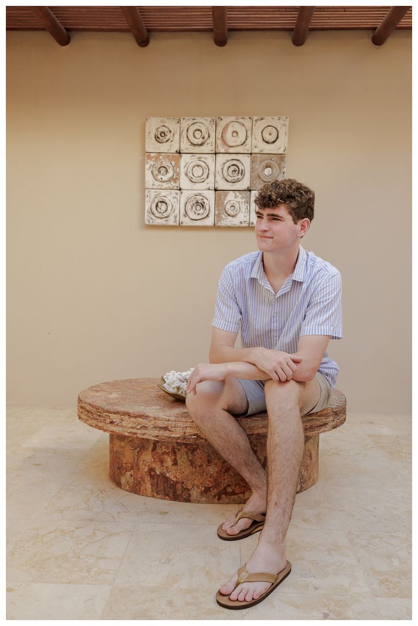 Young man in striped shirt and shorts sitting on a rustic stone bench against a beige wall with decorative tiles.