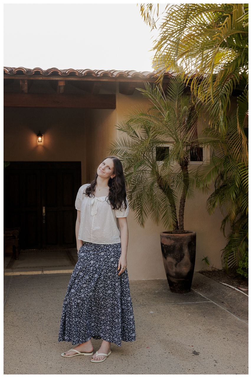 Young woman in white top and floral maxi skirt standing outside a Mediterranean-style building with a palm tree.