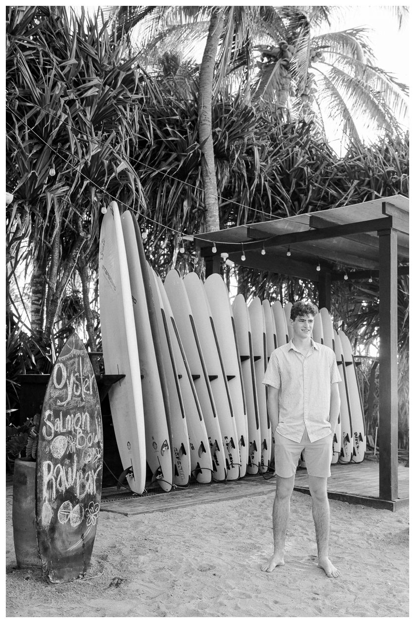 Man standing beside surfboard rack at tropical beach surf shop with palm trees in black and white photo.