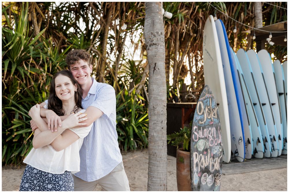 Couple embracing and smiling near surfboards and tropical greenery at a beach location.