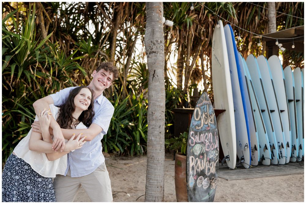 Happy couple embracing and laughing near colorful surfboards leaning against a fence at a tropical beach location.