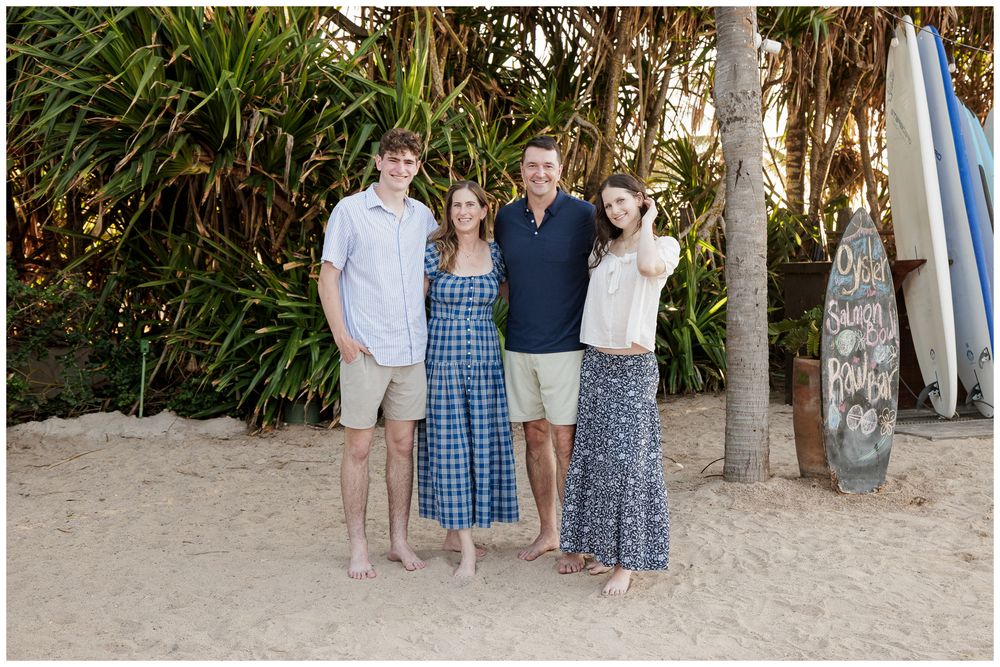 Family of four smiling together on a sandy beach with surfboards and tropical palms in the background.