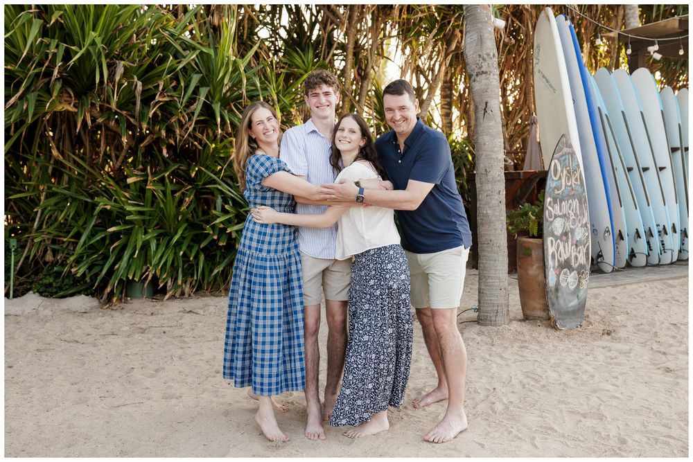 Family of four smiling together on a sandy beach with surfboards and palm trees in the background.