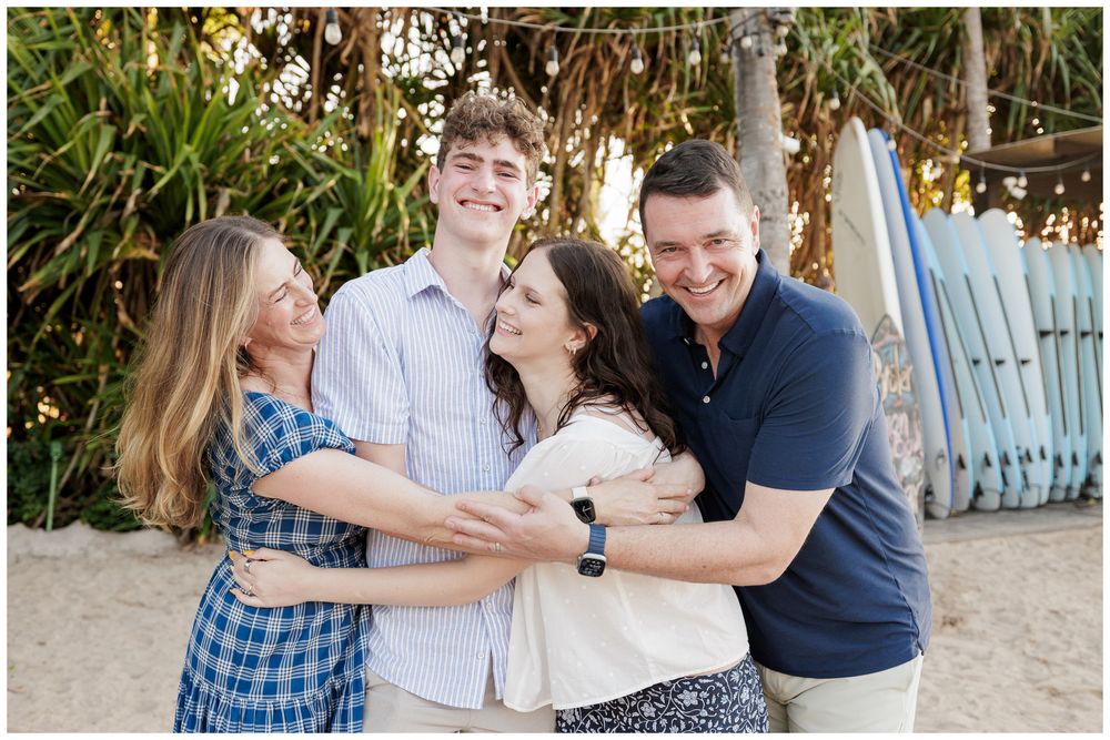 Family of four laughing and hugging together on a beach with colorful surfboards in the background.