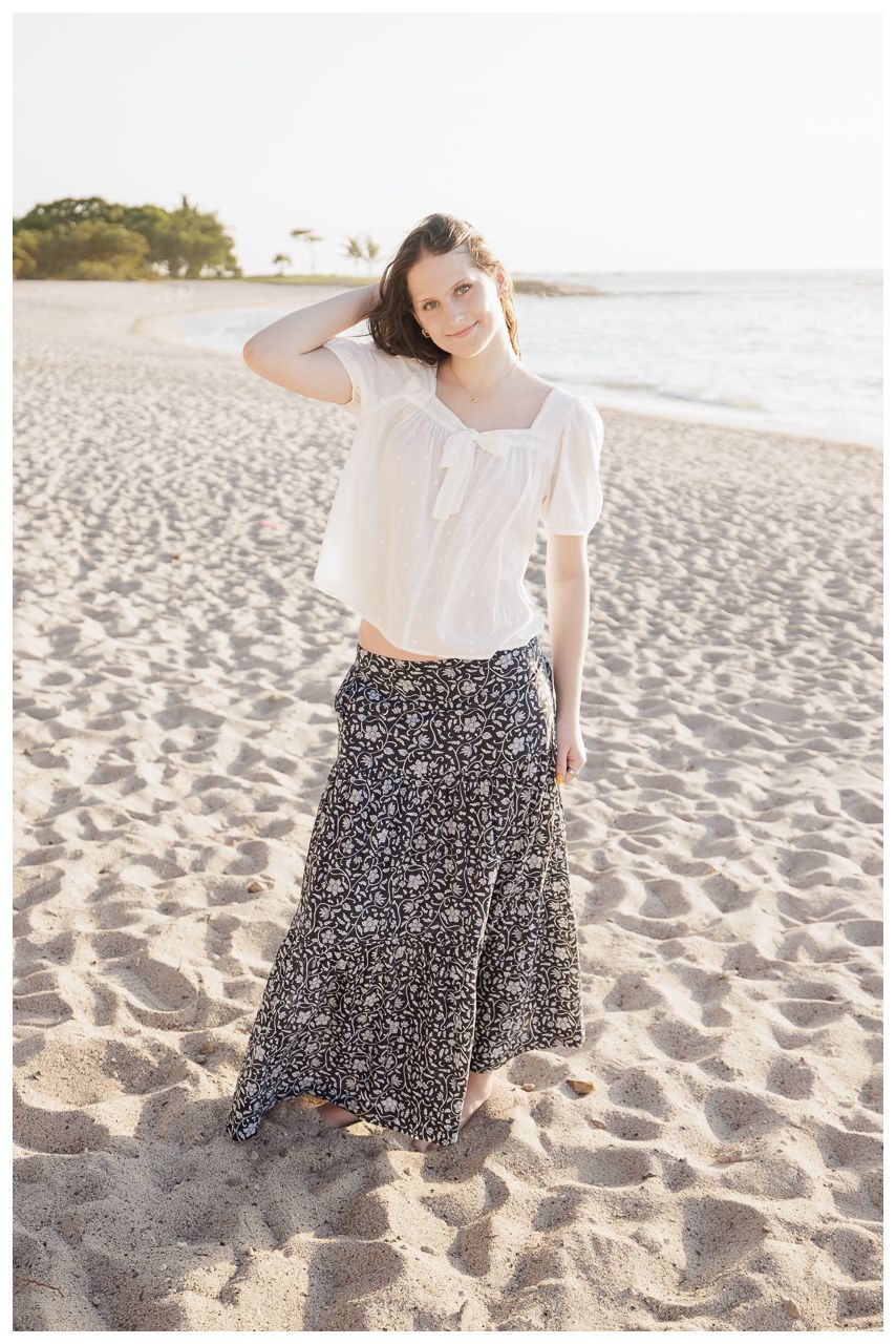 Young woman in white top and floral maxi skirt standing barefoot on sandy beach at golden hour sunset.