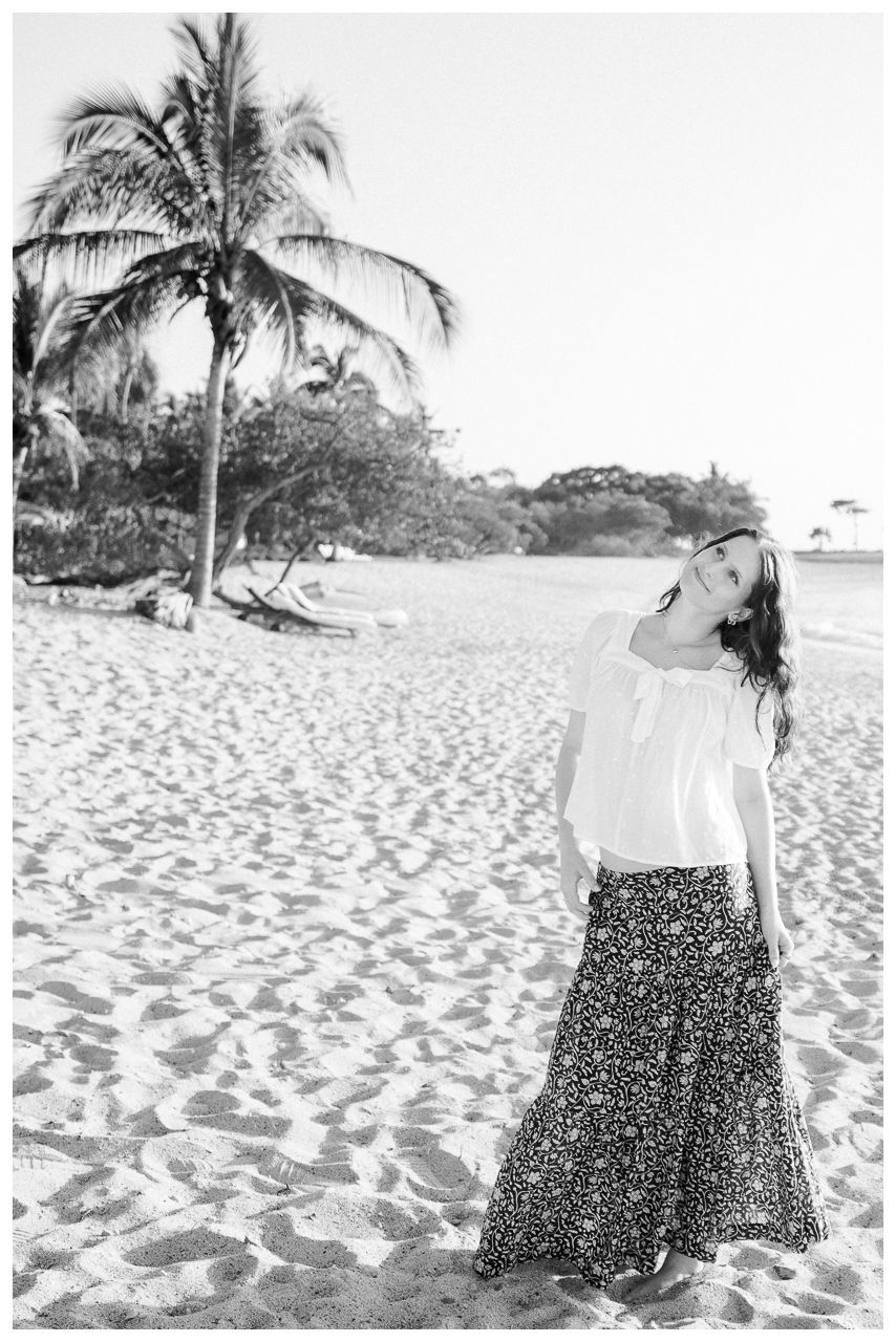 A young woman in a white top and floral maxi skirt laughs joyfully on a sandy beach with a palm tree.