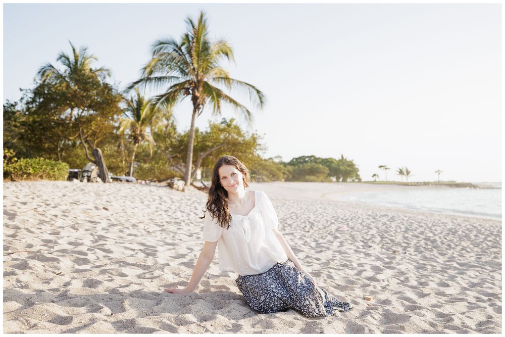 Young woman in white top and floral skirt sitting on white sand beach with palm trees in golden light.