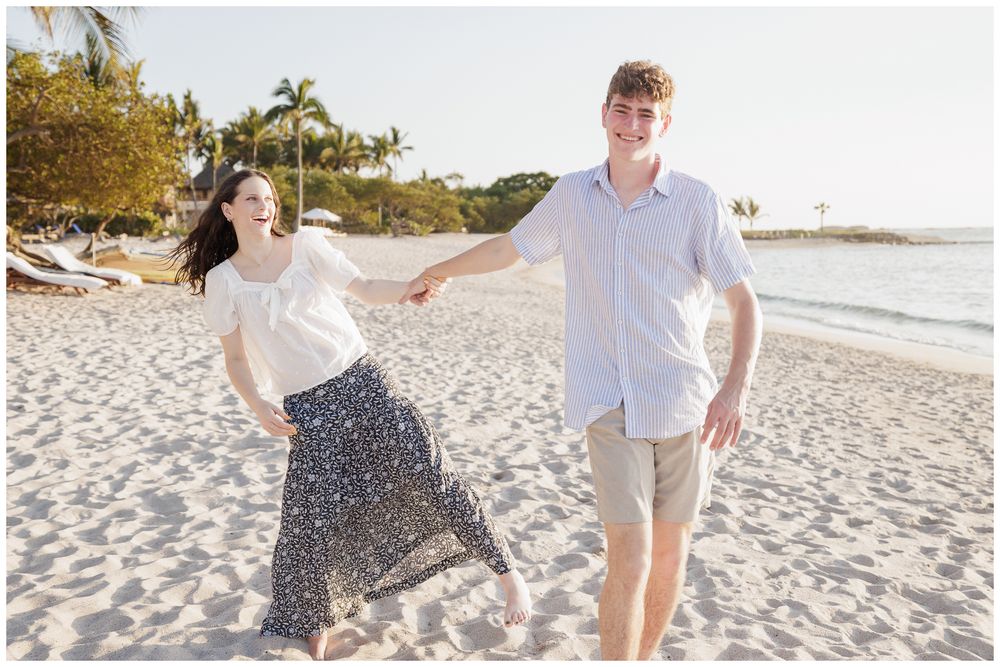Joyful couple holding hands and walking together on a sunny white sand beach with palm trees in background.