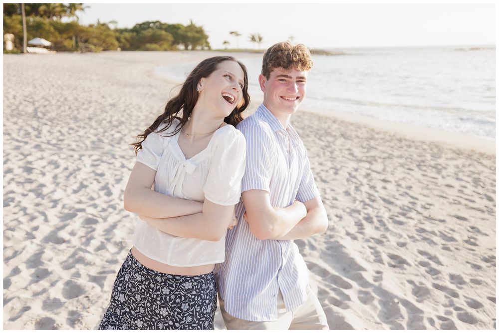 Two teenagers laughing back-to-back on a sunny sandy beach during a family photo session.