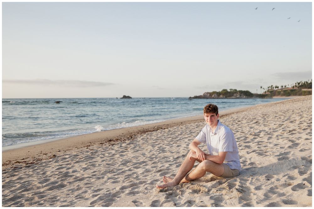 Young man in white shirt and shorts sitting on sandy beach at sunset with ocean waves and tropical shoreline.