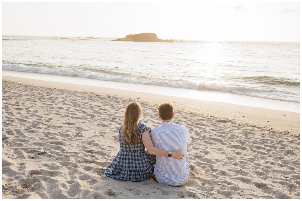 A couple sits together on a sandy beach, embracing while gazing at the ocean at sunset.