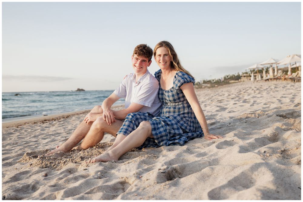 A young couple sits together on a sandy beach at golden hour, smiling warmly in casual blue outfits.