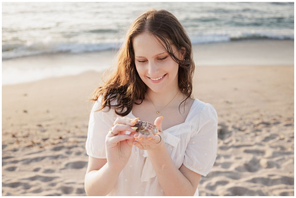 Young woman in white dress smiling at seashell on sandy beach at golden hour.