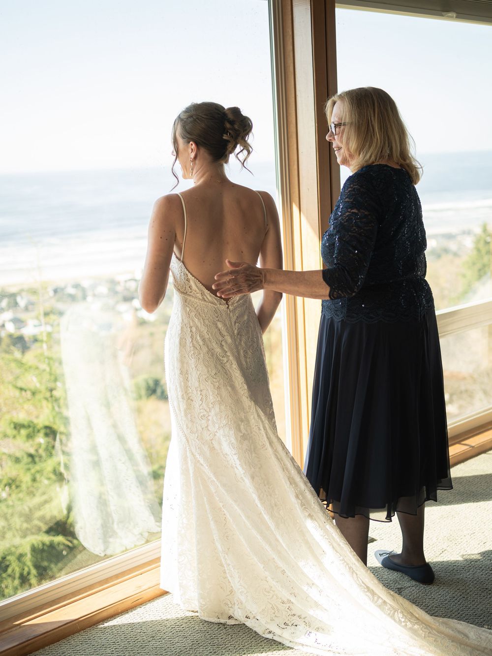 Mother helping bride with backless lace wedding dress near large window with ocean view.